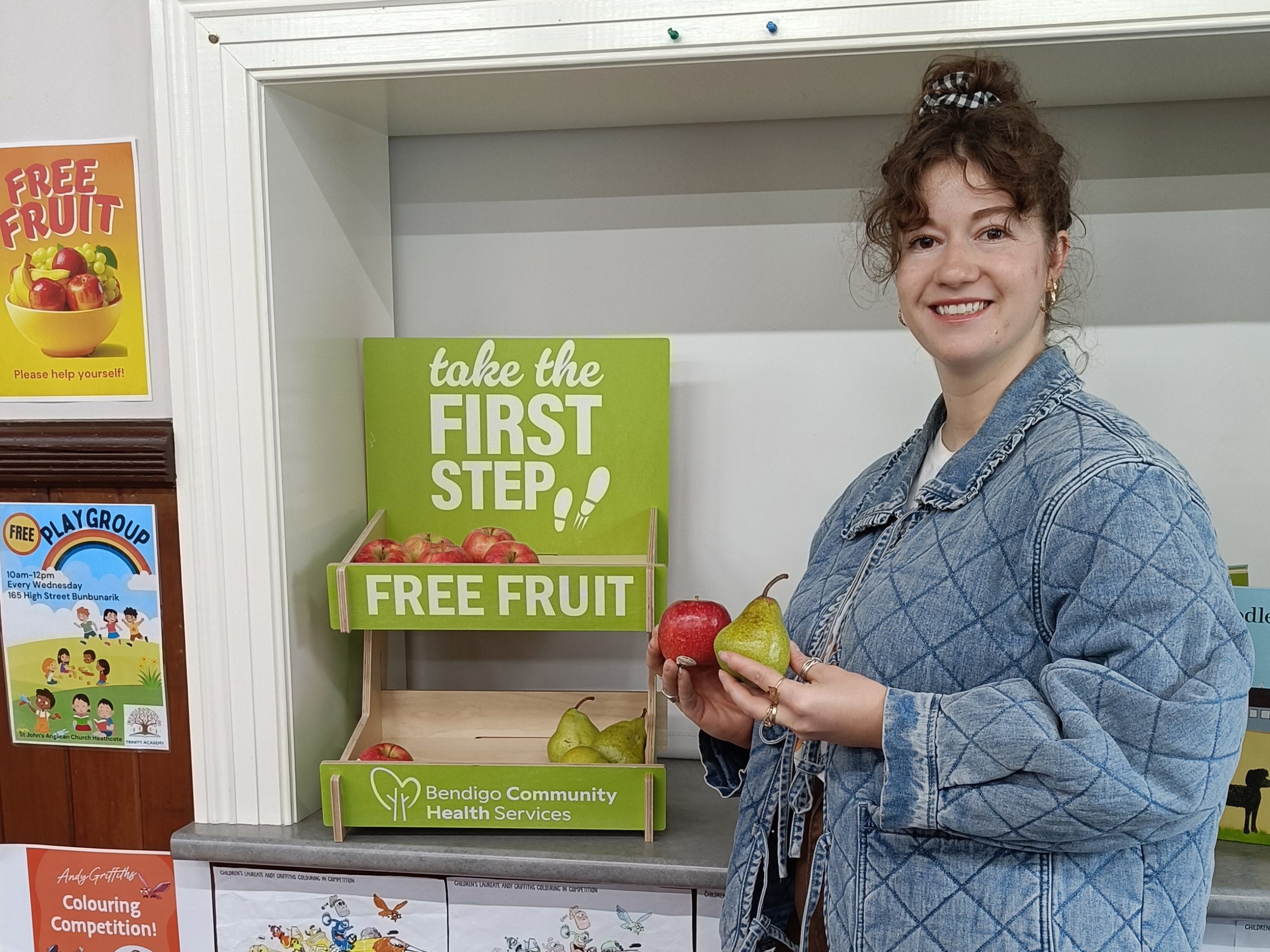 Olivia stands beside a small green fruit stand, holding some fruit.